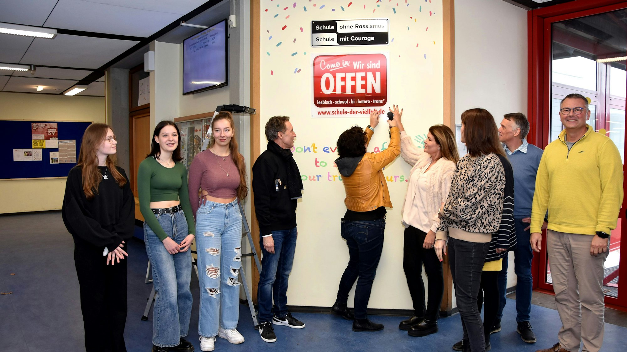 Schülerinnen und Lehrkräfte stehen vor einem Schild in einer Schule.