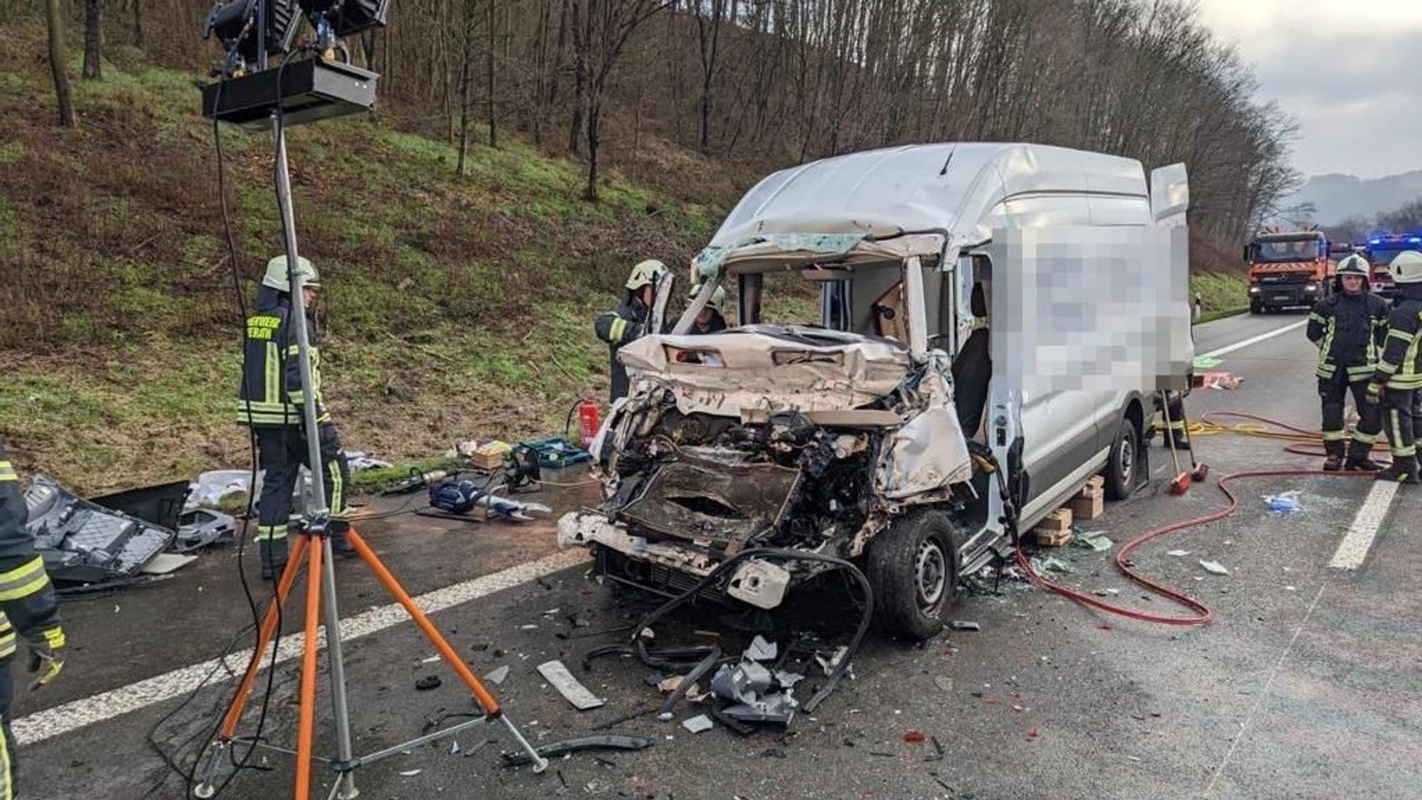 Ein schwer beschädigter Kastenwagen steht auf der Autobahn. Teile des Fahrzeugs liegen auf der Fahrbahn. Feuerwehrleute stehen neben dem Wagen.