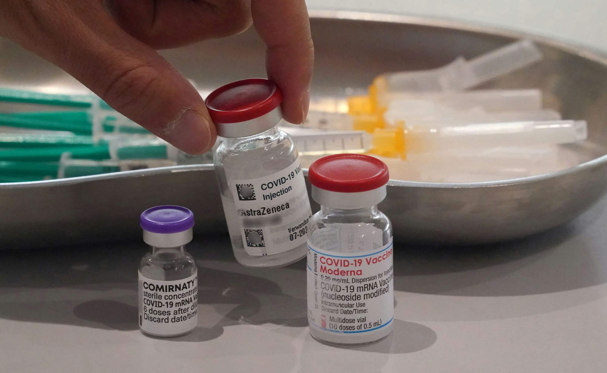 Ein Mitarbeiter eines Impfzentrums nimmt eine Impfdose vom Impfstoff Astrazeneca vom Tisch. Links im Bild steht eine Impfdose vom Impfstoff Biontech, rechts im Bild steht eine Impfdose vom Impfstoff Moderna.