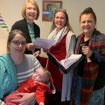 Judith Wittköpper sitzt am Tisch, ihr Baby im Arm, die anderen Frauen stehen hinter ihr. Sie lächeln in die Kamera.