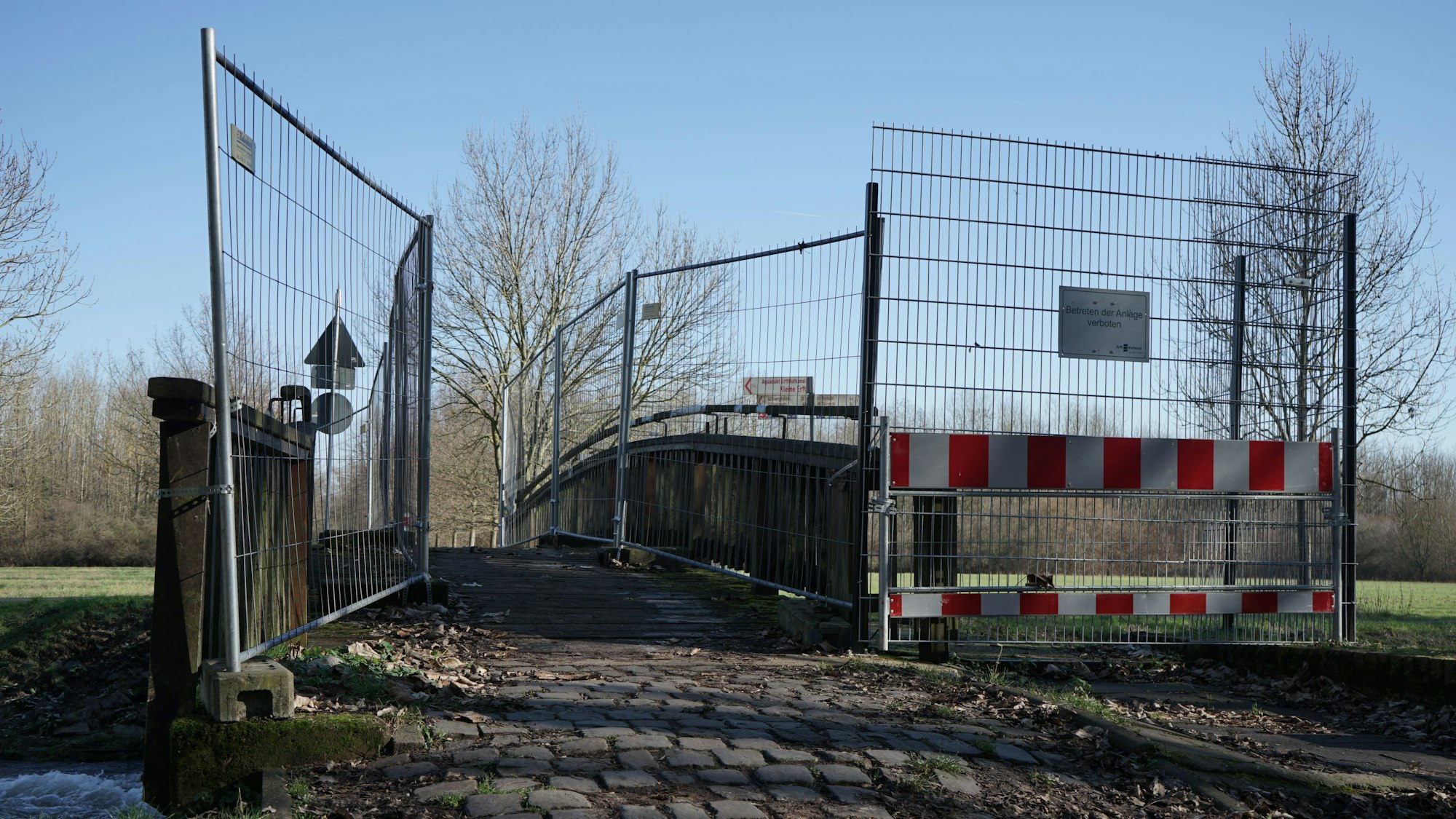 Man sieht eine schmale Brücke über ein Gewässer, welche an den Geländern durch Bauzäune begrenzt wird.