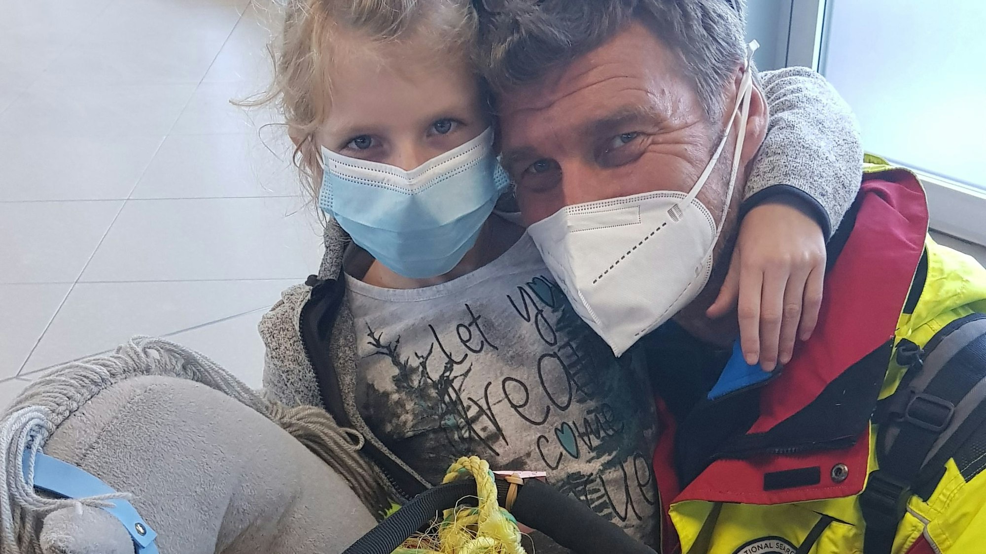 man sihet Vater und Tochter, wie sie in die Kamera gucken, sie tragen Masken. Ingo hat sich neben Mia gekniet, diese hat den Arm um seinen Hals.