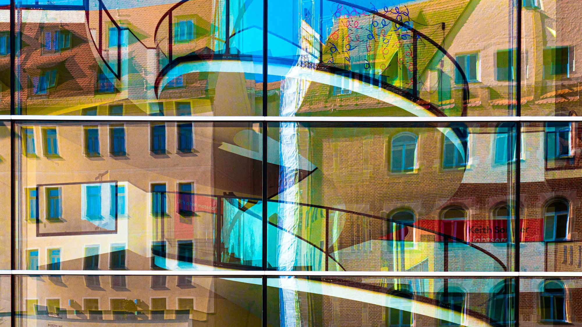 Hausfassaden spiegeln sich verzerrt und bunt in einer Glasfront, hinter der eine Wendeltreppe durchscheint.