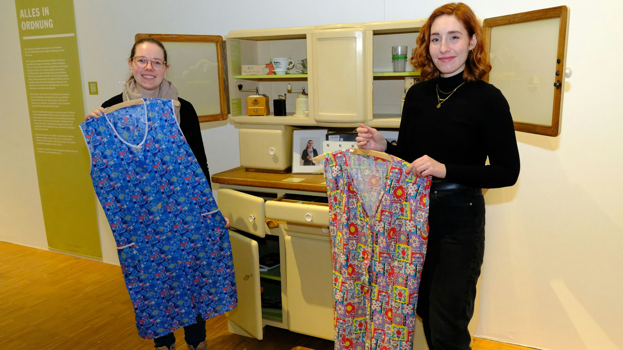Ann Heinen (re.) und Hannah Drießler mit Kittelschürzen vor einem Buffetschrank in der Ausstellung Herdanziehungskraft im LVR-Freiliichtmuseum Kommern.