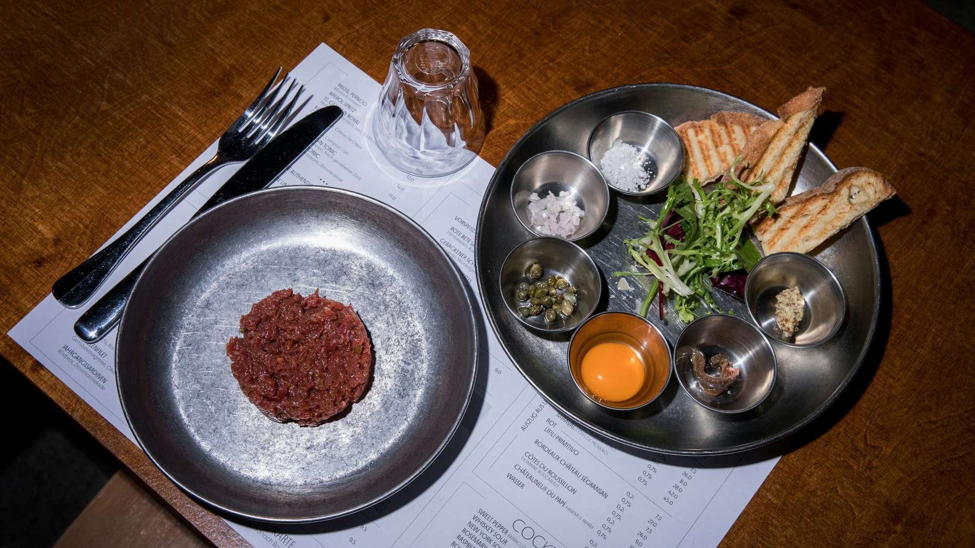 Rindertartar auf einem Teller, daneben ein Teller mit geröstetem Brot und kleinen Schälchen mit Salz, Kapern und Eigelb