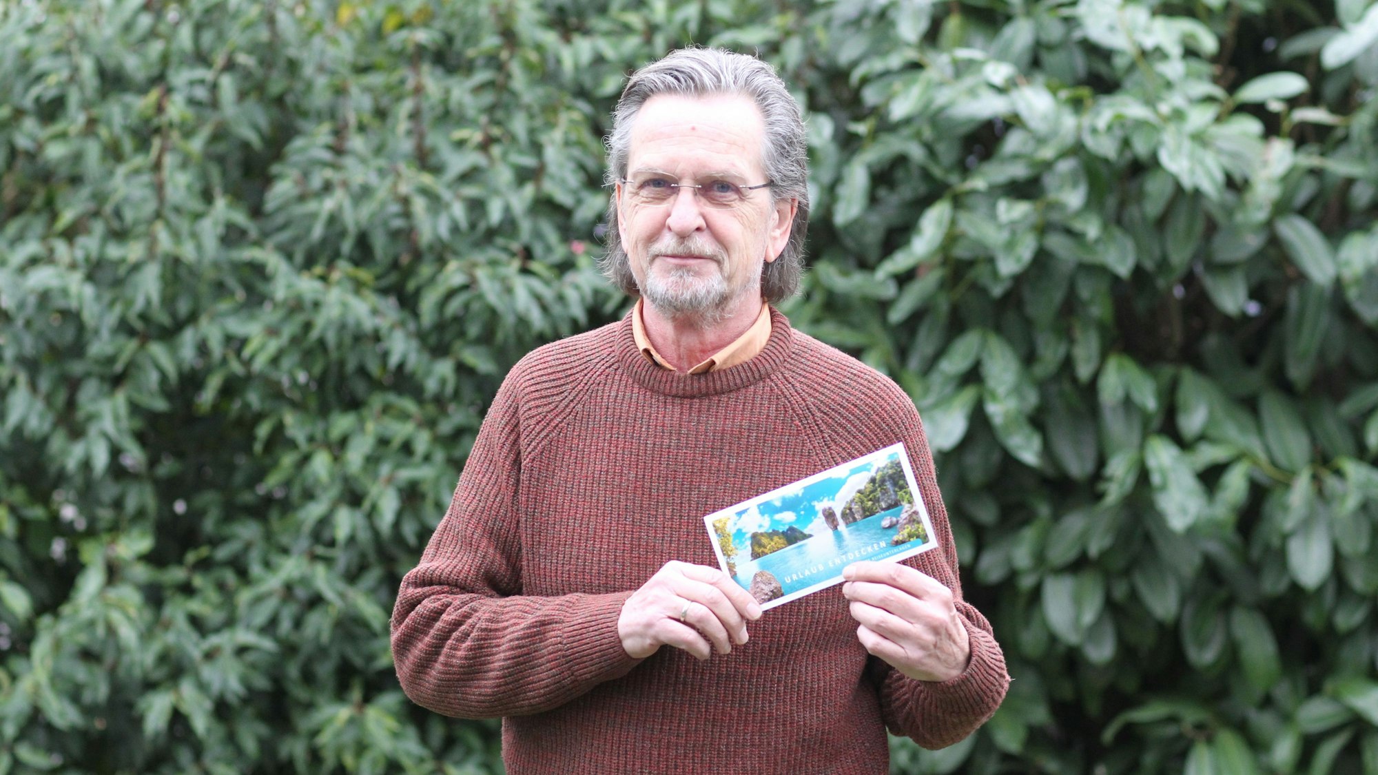 Ein bebrillter Mann mit grauen Haaren und Bart steht vor einer Hecke. Er trägt einen Pullover und hält ein Karte in den Händen, die ein Urlaubsparadies zeigt.