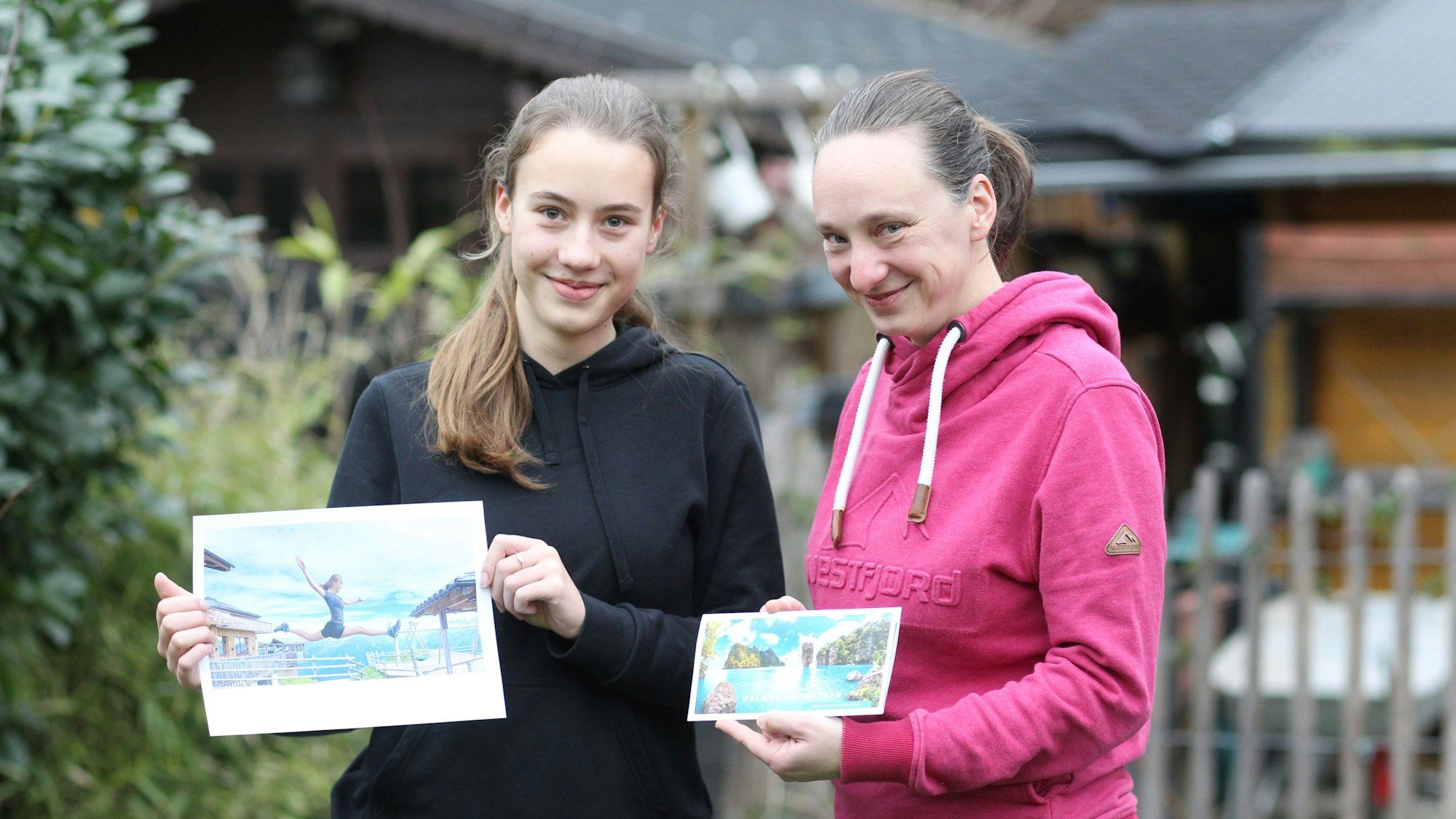 Eine junge Frau mit Pferdeschwanz hält das Foto von ihr beim Spagatsprung in die Höhe. Neben ihr steht eine weitere Frau, ebenfalls mit Pferdeschwanz, in einem pinkfarbenen Hoodie, den den Reisegutschein in der Hand hält.