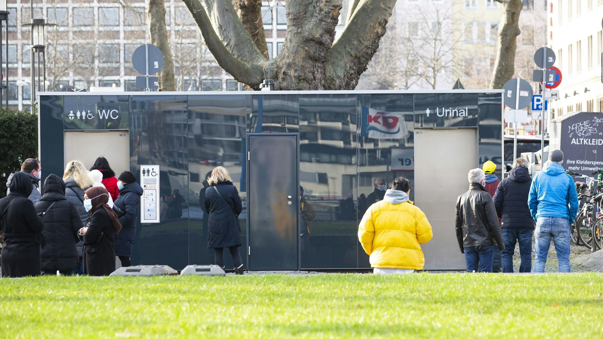Vor einer öffentlichen Toilette am Kölner Rheinufer stehen Personen an.