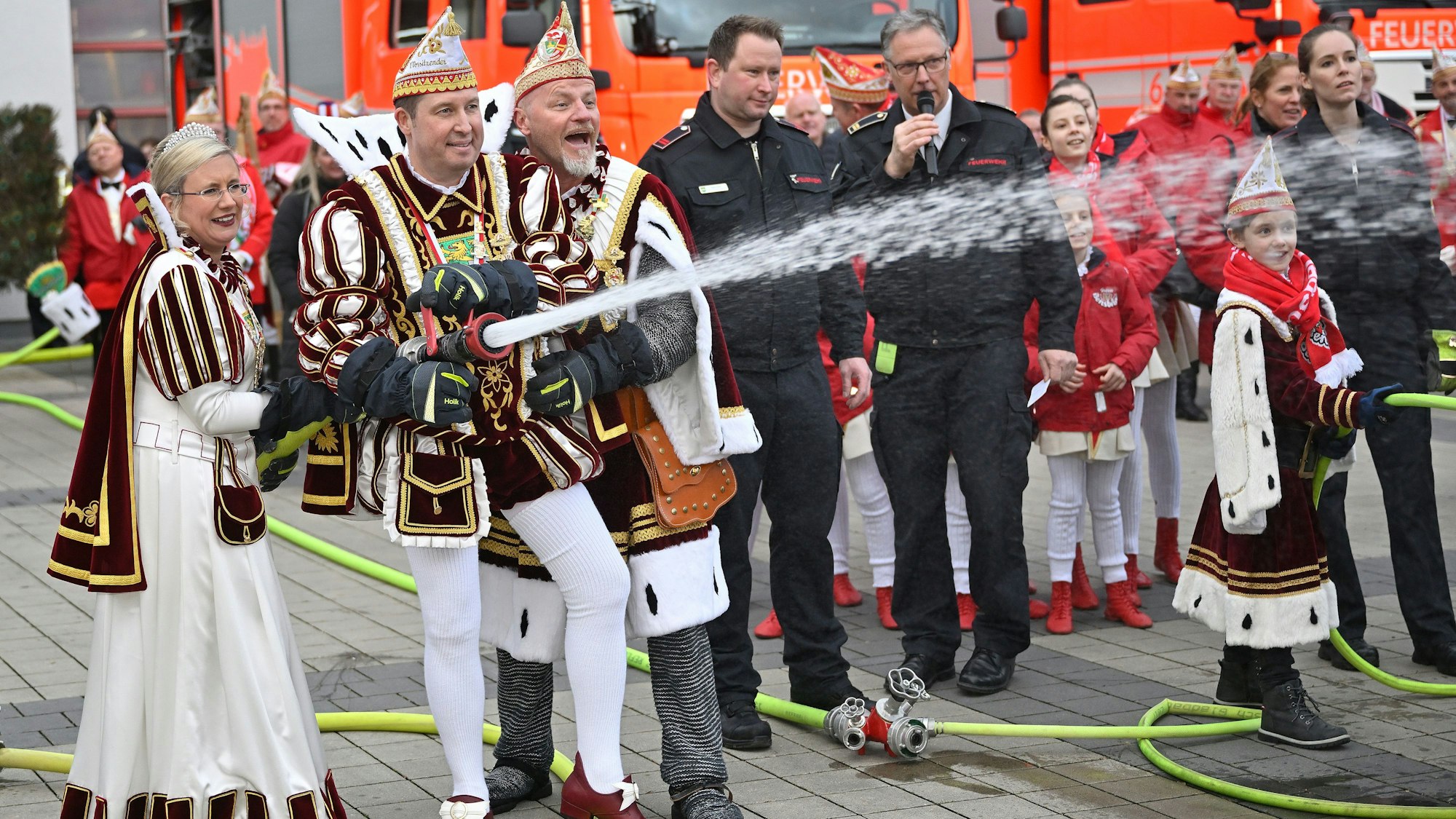 Prinz, Bauer und Jungfrau halten ein Feuerwehrstrahlrohr aus dem Wasser spritzt. Im Hintergrund sind Feuerwehrfahrzeuge zu sehen.