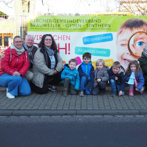 Ein Plakat mit der Aufschrift „Wir brauchen Dich!“. Kita-Mitarbeiter und Kinder stehen davor.