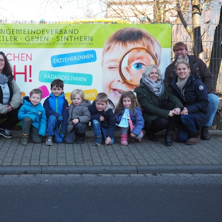 Ein Plakat mit der Aufschrift „Wir brauchen Dich!“. Kita-Mitarbeiter und Kinder stehen davor.
