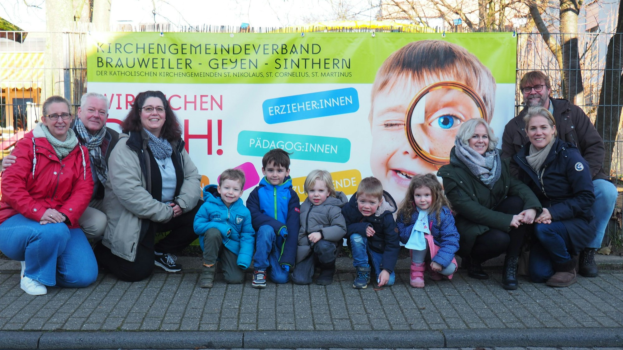 Ein Plakat mit der Aufschrift „Wir brauchen Dich!“. Kita-Mitarbeiter und Kinder stehen davor.