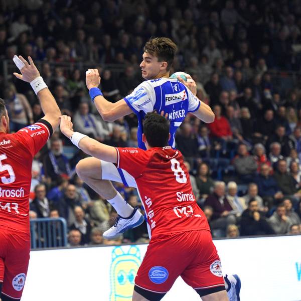 Mehrere Handballspieler zweier Mannschaften sind in einer Spielsituation zu sehen. Einer setzt mit Ball in der Hand zum Sprungwurf an.