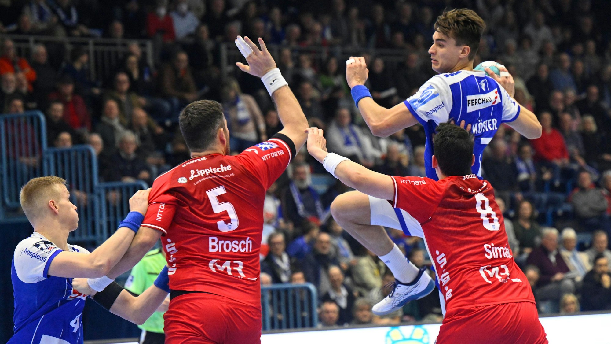 Mehrere Handballspieler zweier Mannschaften sind in einer Spielsituation zu sehen. Einer setzt mit Ball in der Hand zum Sprungwurf an.