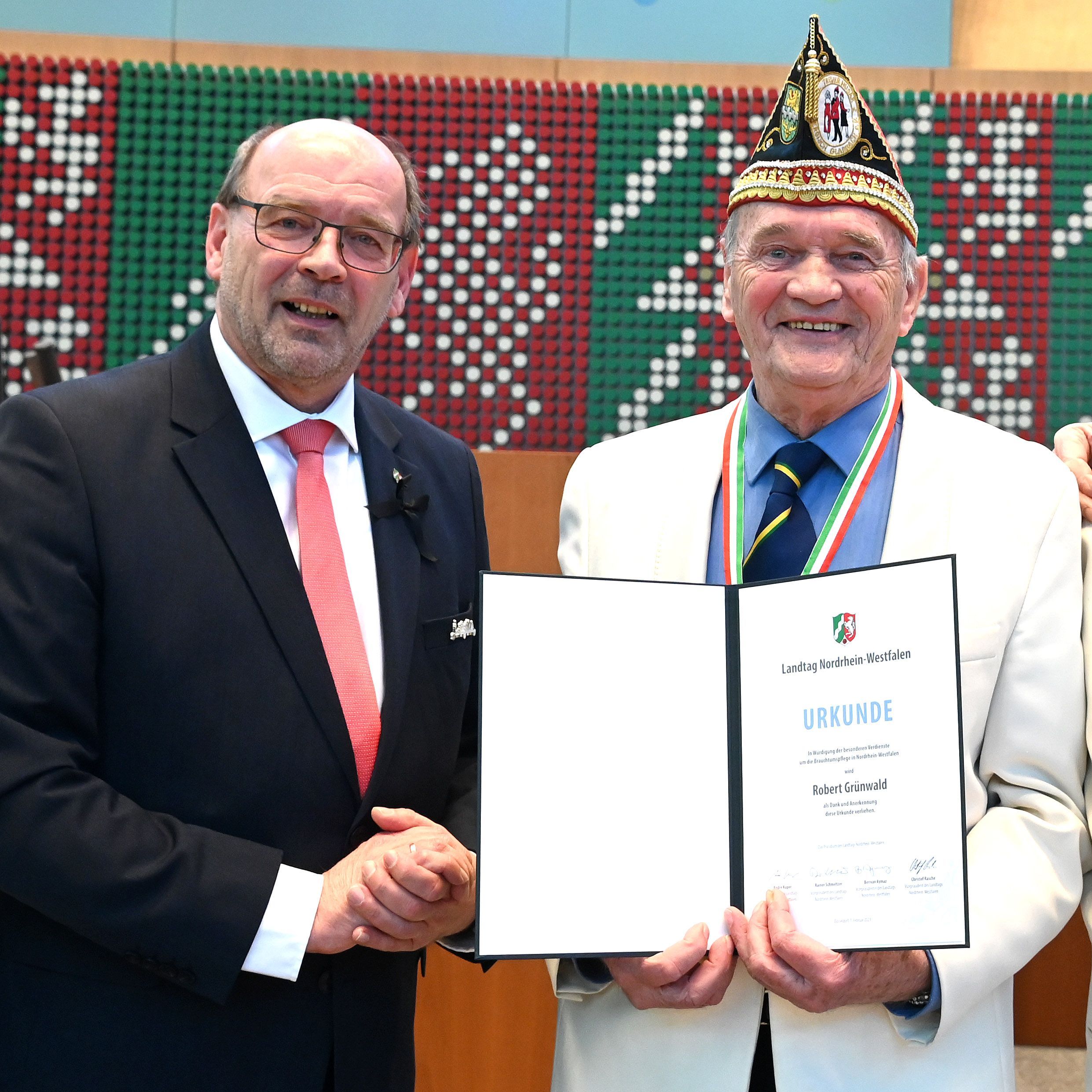 Es sind Rainer Schmeltzer und Robert Grünwald zu sehen. Grünwald hält eine Urkunde in den Händen.