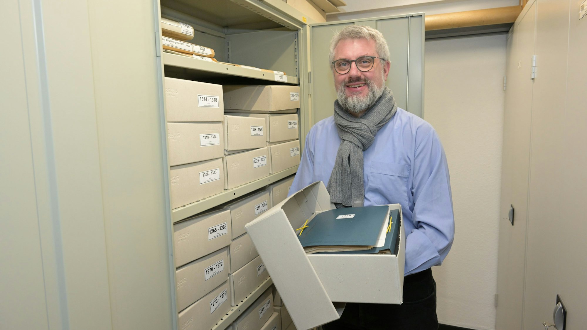 Es ist der Pfarrer zu sehen, der eine Kiste mit Dokumenten in den Händen hält.