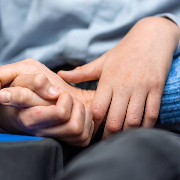 Die Hände eines Kindes, das im Rollstuhl sitzt, halten eine Hand seiner Mutter fest.