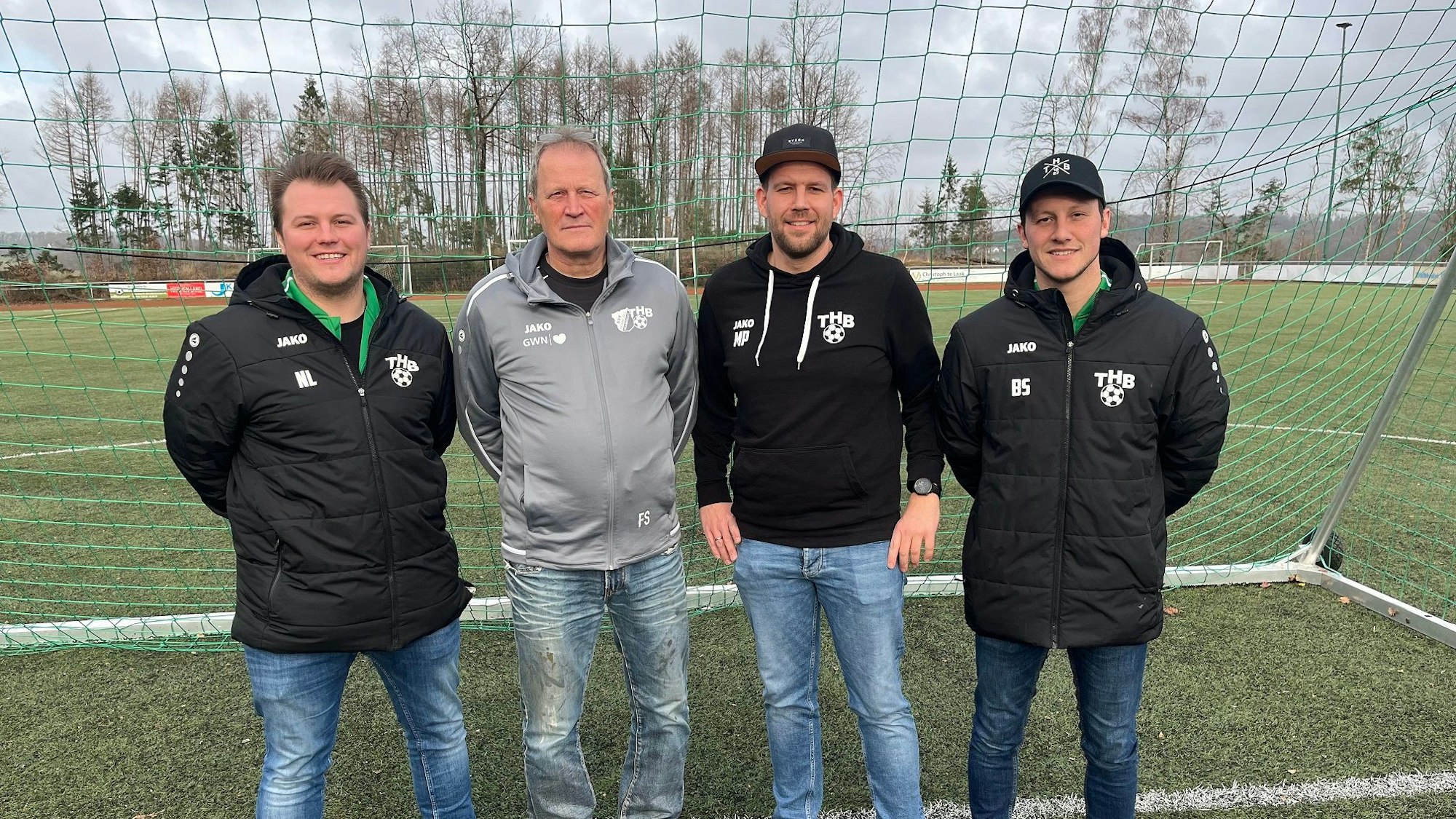 Vier Männer stehen in sportlicher Kleidung nebeneinander in einem Tor auf einem Fußballplatz.