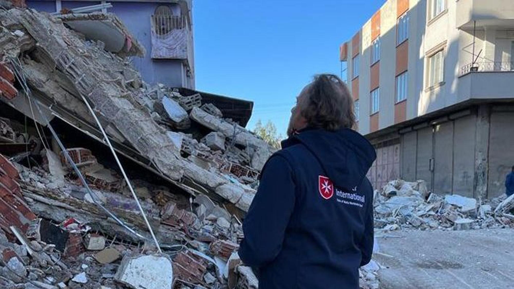 Eine Frau mit einer „Malteser International“-Jacke steht vor einem Trümmerhaufen.