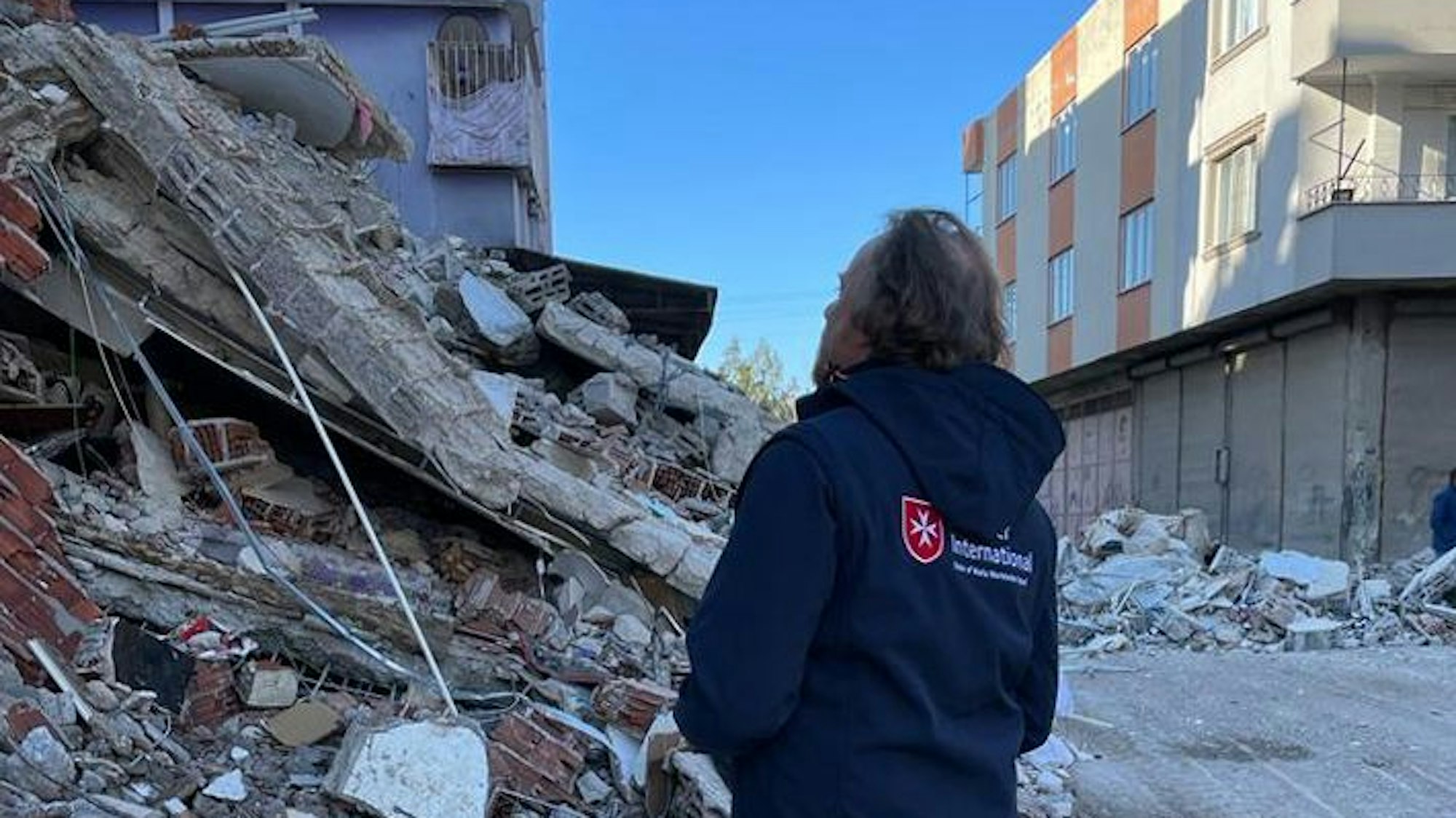 Eine Frau mit einer „Malteser International“-Jacke steht vor einem Trümmerhaufen.