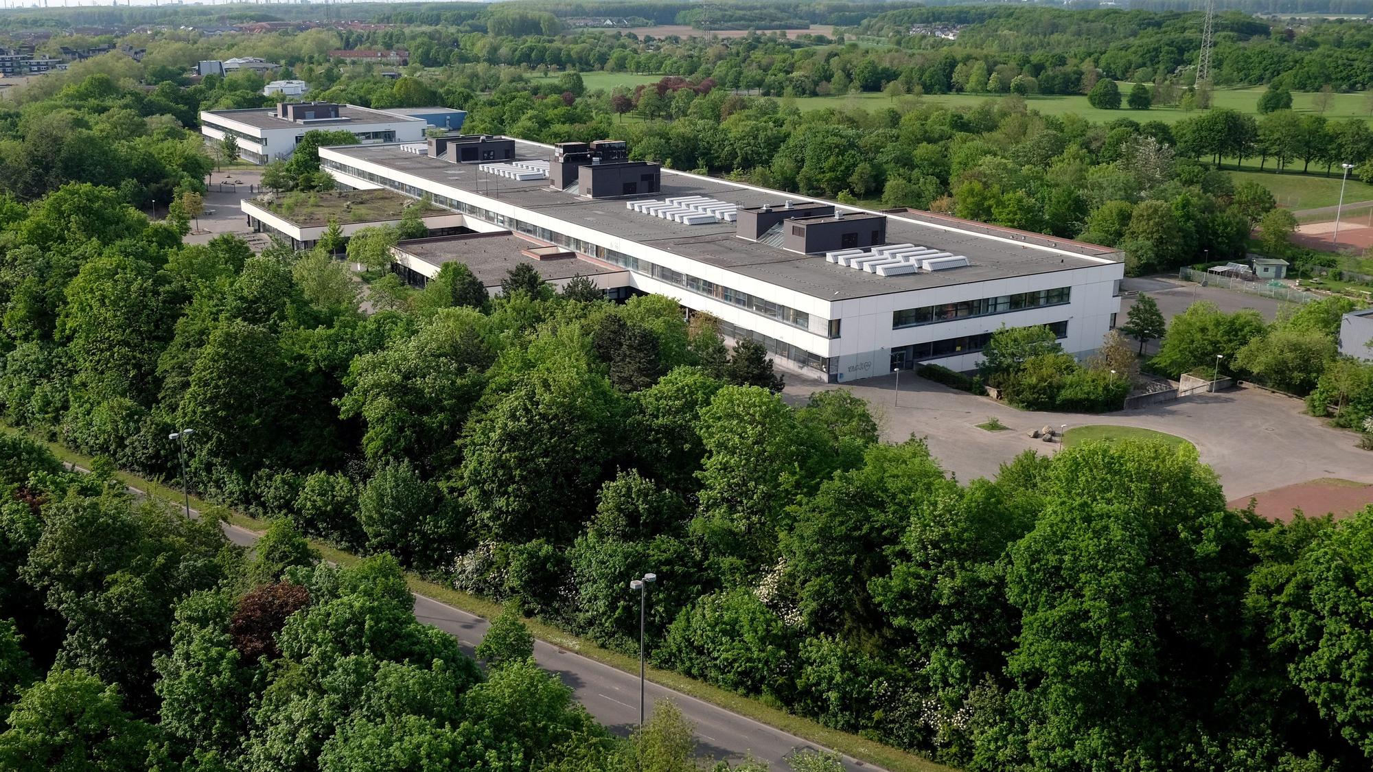 Ein langgezogenes zweistöckiges Gebäude mit Flachdach ist von oben zu sehen, drumherum sind Bäume und Wiesen und eine Straße.