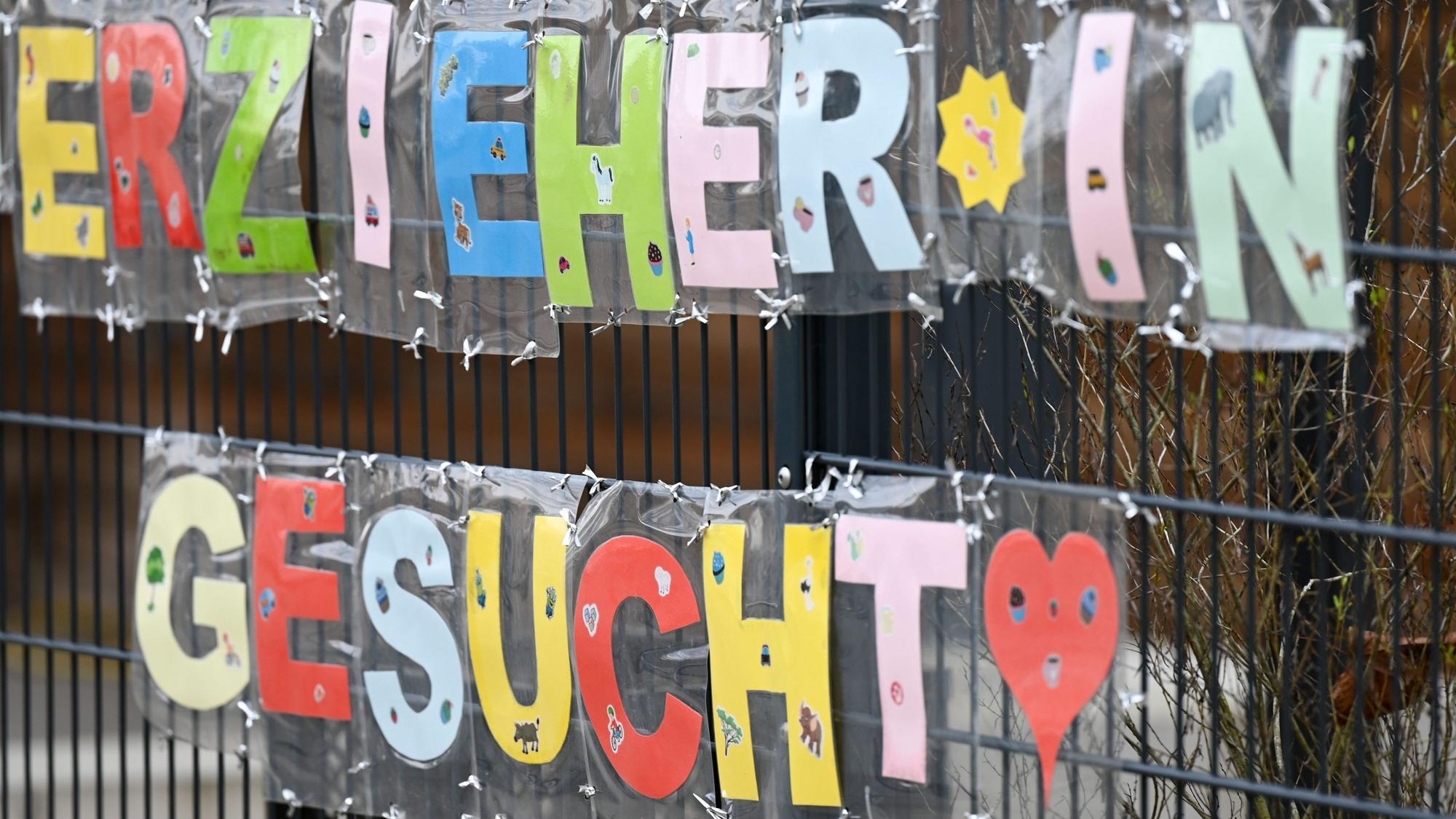 Man sieht ein großes Transparent an einem Zaun hängen, auf ihm steht „Erzieher*in gesucht“.