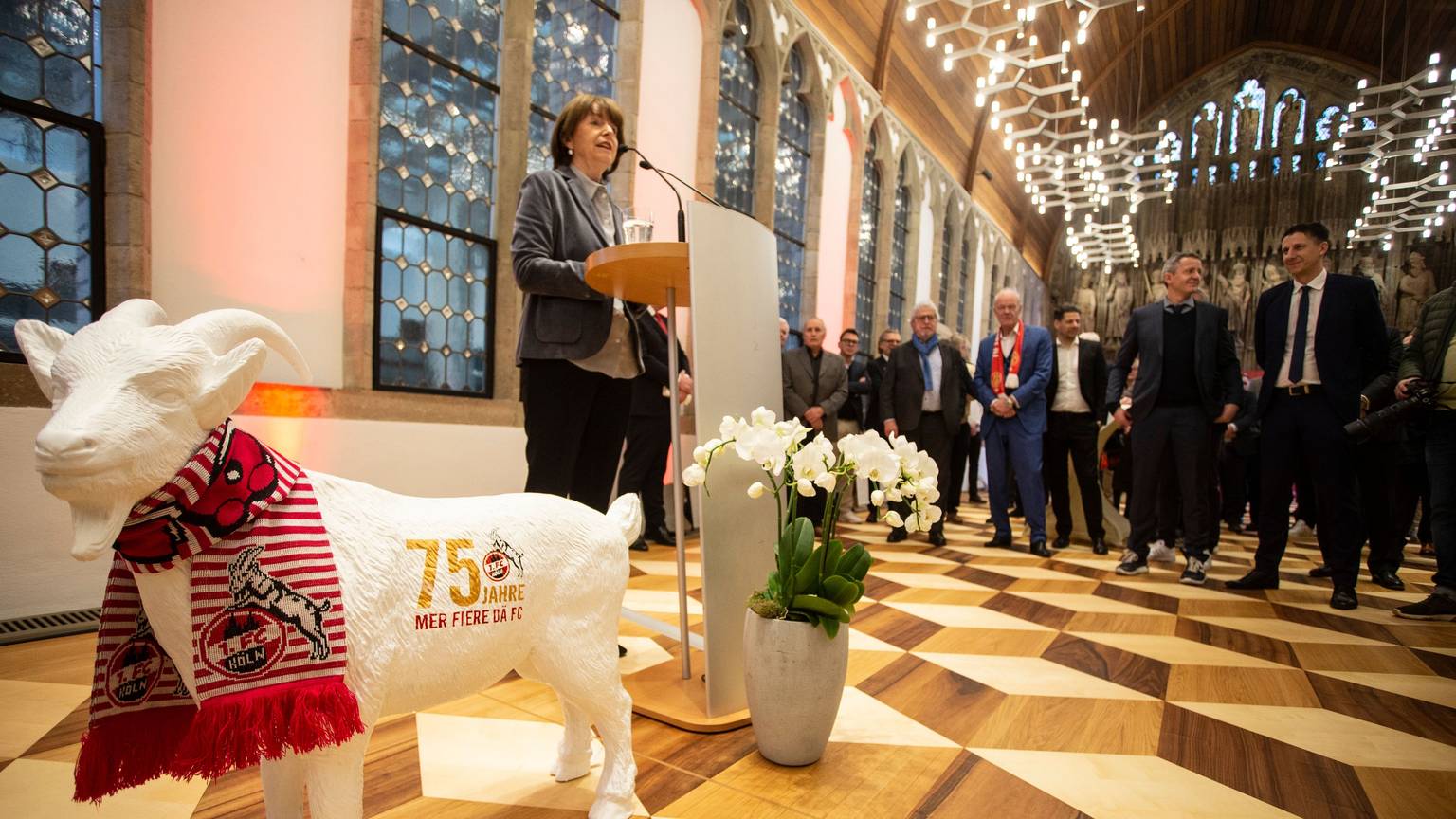 Die Kölner Oberbürgermeisterin Henreitte Reker steht im Historischen Rathaus an einem Rednerpult. Im Vordergrund steht eine Geißbock-Skulptur, im Hintergrund einige FC-Legenden.