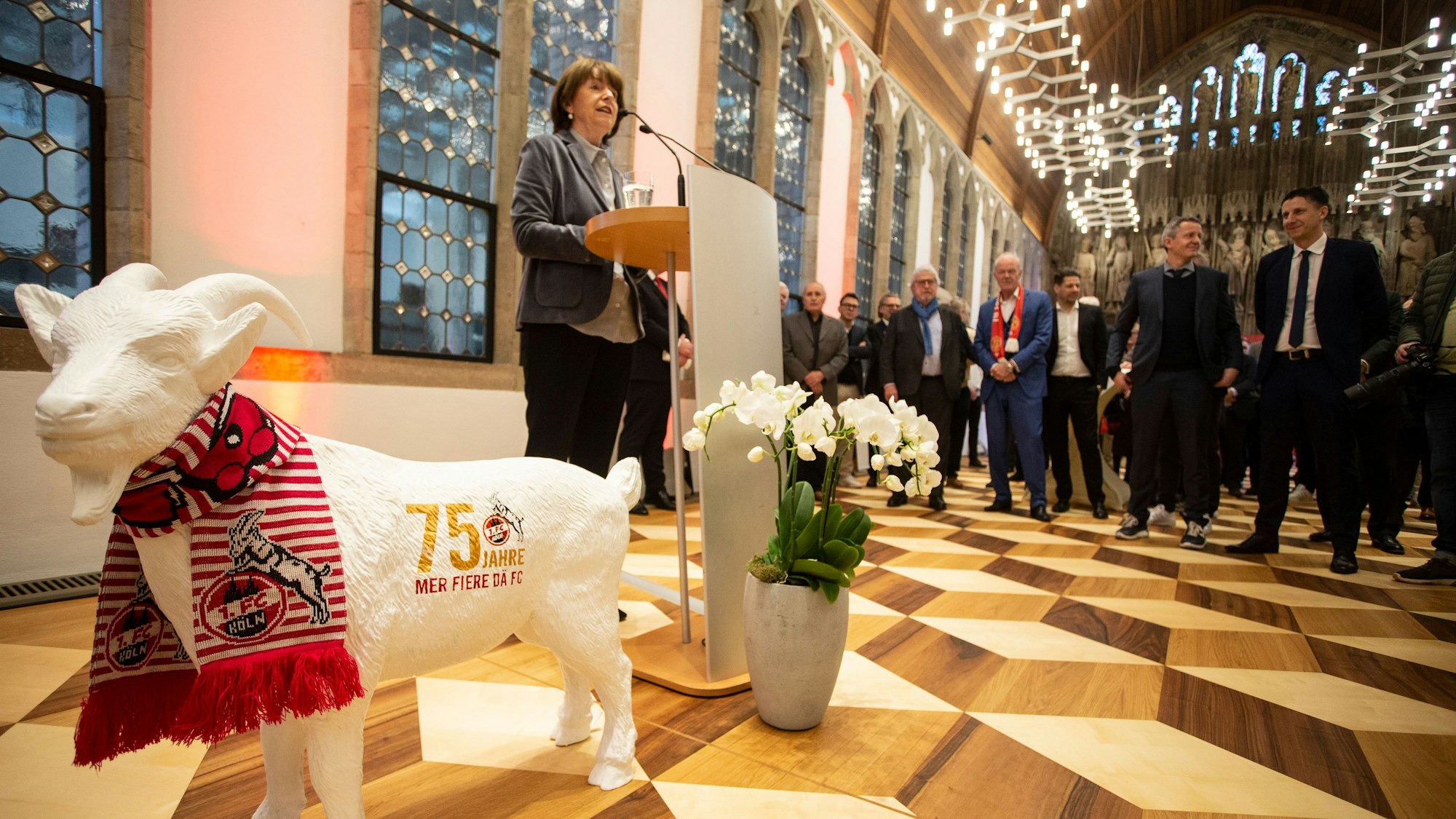 Die Kölner Oberbürgermeisterin Henreitte Reker steht im Historischen Rathaus an einem Rednerpult. Im Vordergrund steht eine Geißbock-Skulptur, im Hintergrund einige FC-Legenden.