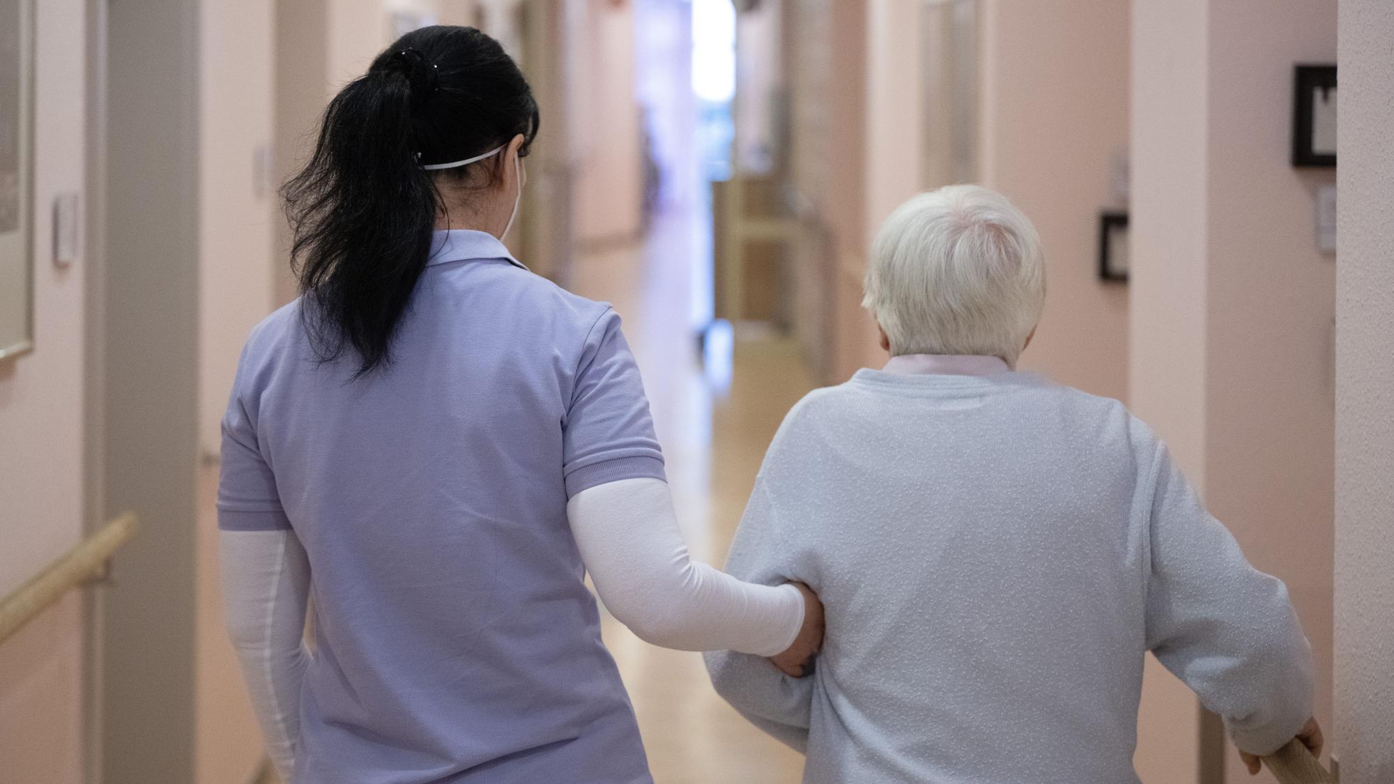 Eine Pflegefachkraft geht mit einer alten Frau über den Gang eines Pflegeheims.