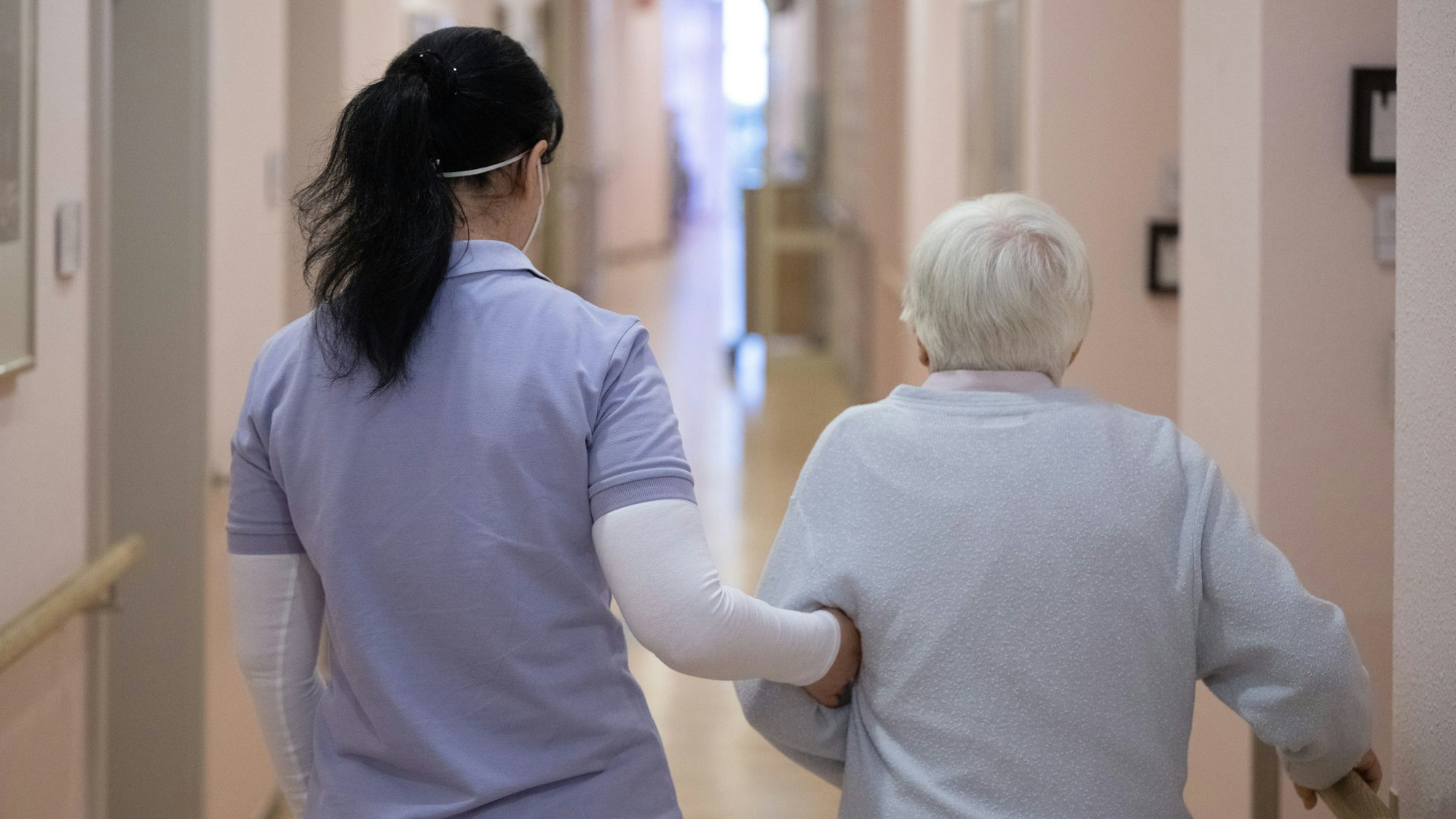 Eine Pflegefachkraft geht mit einer alten Frau über den Gang eines Pflegeheims.