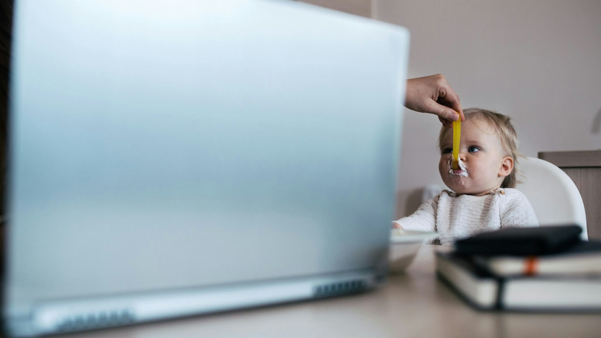 Eine Hand kommt hinter dem Laptop hervor und füttert ein Baby.