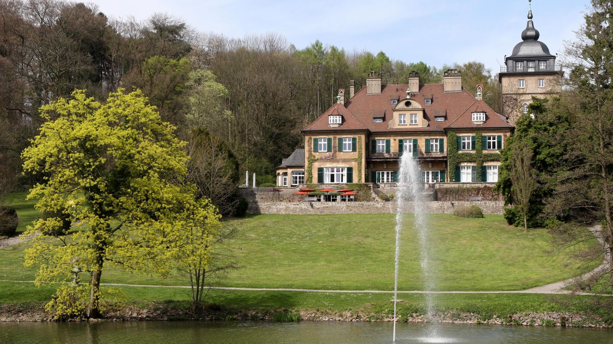 Hinter einem Teich mit Fontäne steht oberhalb einer Wiese ein herrschaftliches Haus mit Turm.