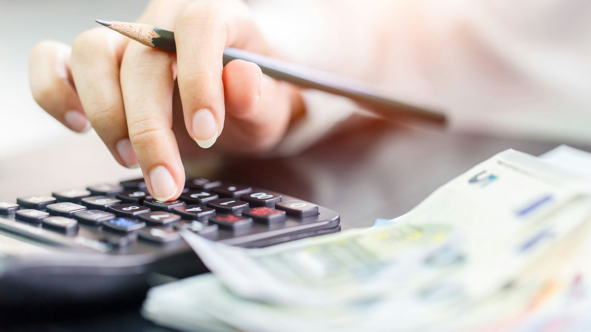 Eine Frau hält einen Bleistift in einer Hand und tippt auf einem Taschenrechner, daneben liegen Geldscheine.