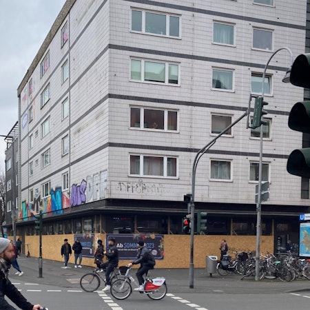 Das Eckhaus der geplanten Aldi-Süd-Filiale ist bisher noch mit Brettern verkleidet, im Vordergrund fahren Fahrradfahrer auf der Straße.