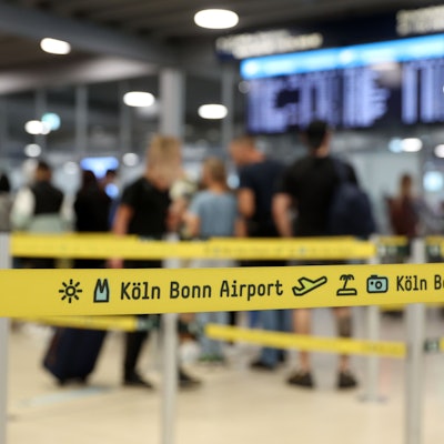 Ein gelbes Absperrband am Flughafen Köln/Bonn. Im Hintergrund sind wartende Passagiere an der Sicherheitskontrolle zu sehen.
