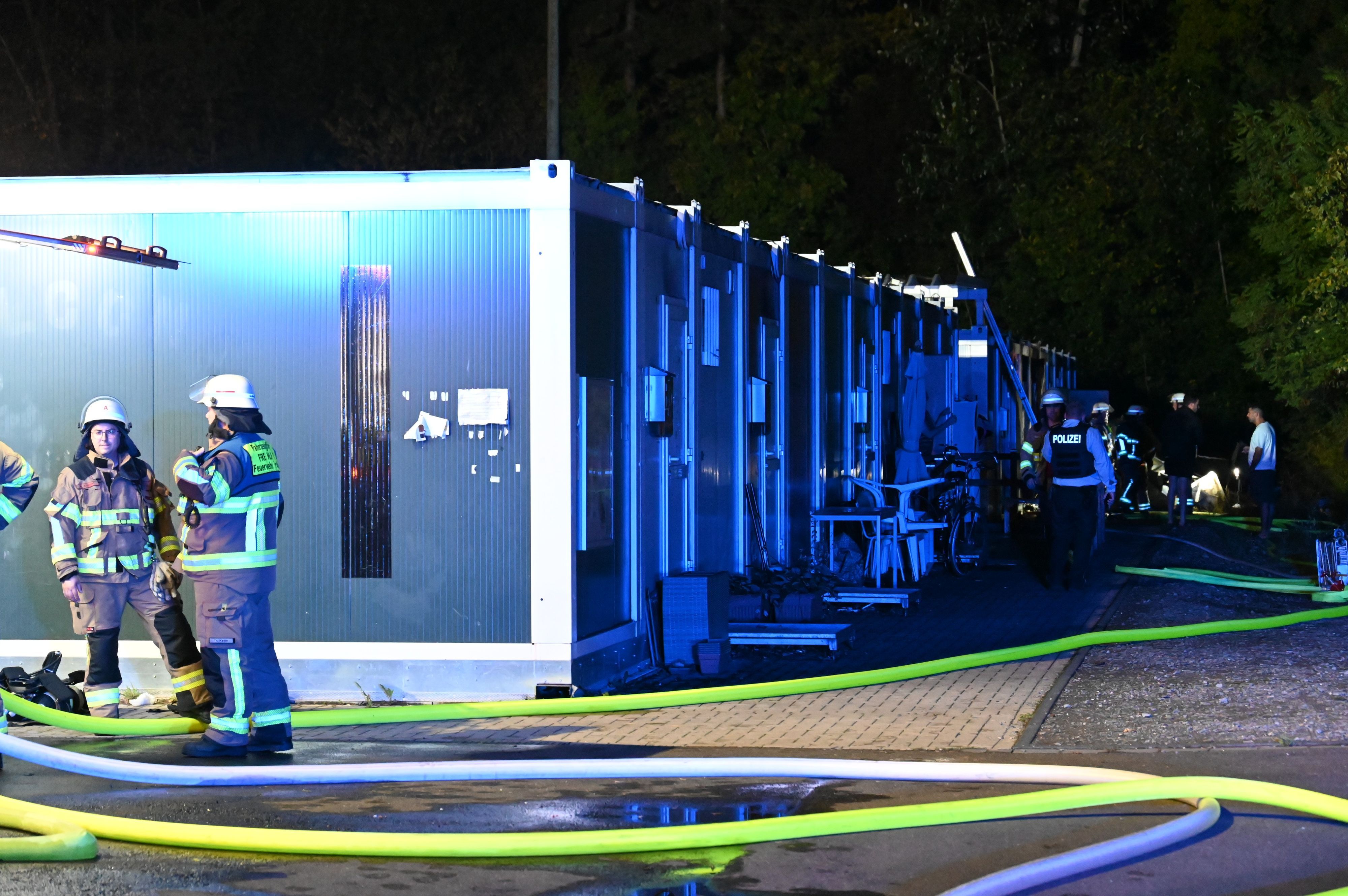 Das Foto zeigt Feuerwehrleute, die vor den Wohncontainern stehen. Der Einsatzort ist hell erleuchtet.