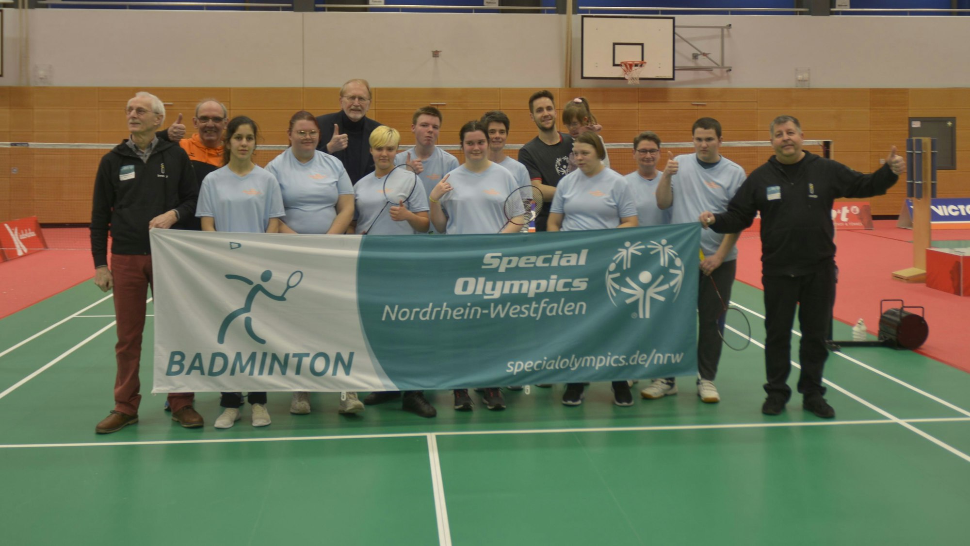Man sieht das Team, wie es in einer Turnhalle hinter dem Teambanner steht und es zusammen hochhält.