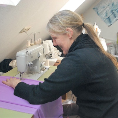 Das Foto zeigt Bekleidungsgestalterin Ursula Fischer. An ihrer Nähmaschine schneidert sie aus lila Stoff ein Kostüm.