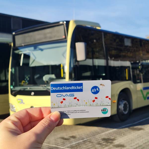 Eine Hand hält ein Deutschlandticket. Im Hintergrund ist ein Bus zu sehen.