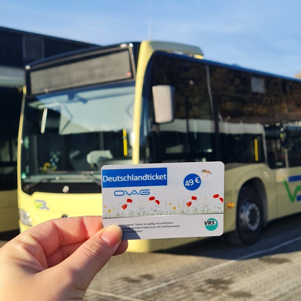 Eine Hand hält ein Deutschlandticket. Im Hintergrund ist ein Bus zu sehen.