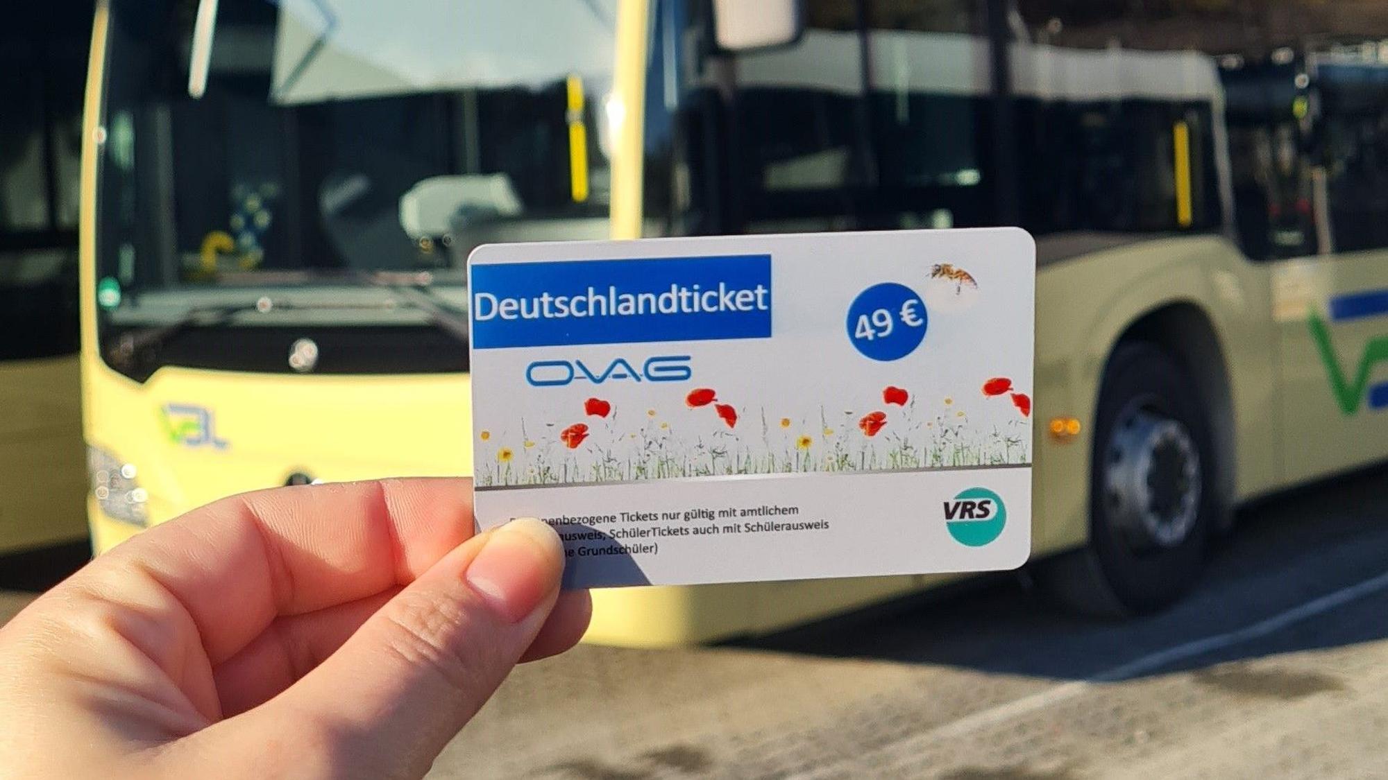 Eine Hand hält ein Deutschlandticket. Im Hintergrund ist ein Bus zu sehen.
