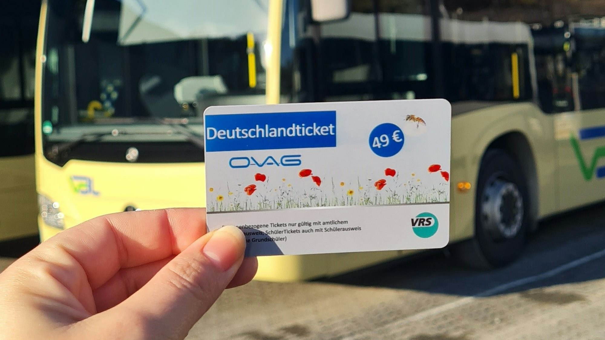 Eine Hand hält ein Deutschlandticket. Im Hintergrund ist ein Bus zu sehen.