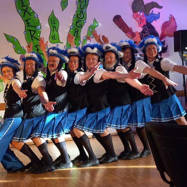Eine Tanzgruppe steht auf einer Bühne in einer Reihe hintereinander und schaut mit ausgebreiteten Armen in Richtung des Publikums.