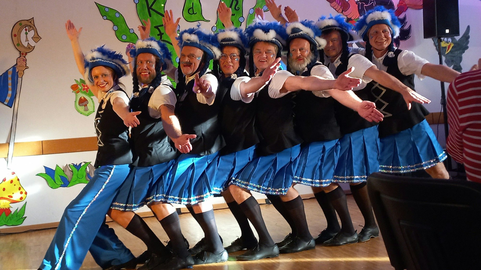 Eine Tanzgruppe steht auf einer Bühne in einer Reihe hintereinander und schaut mit ausgebreiteten Armen in Richtung des Publikums.