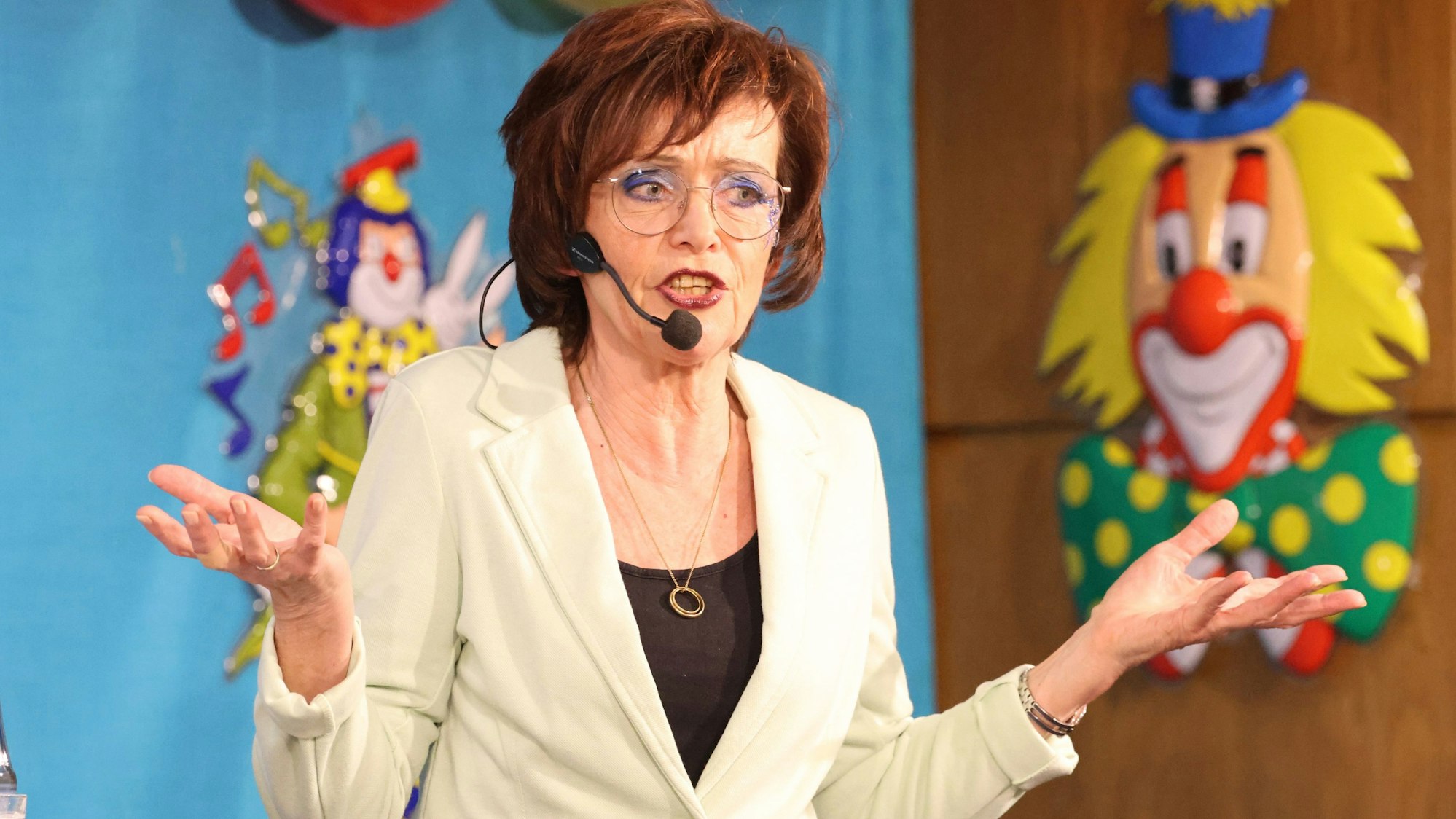 Eine Frau mit einem tragbaren Headset-Mikrofon steht auf einer Bühne, erzählt und breitet dabei die Arme aus.