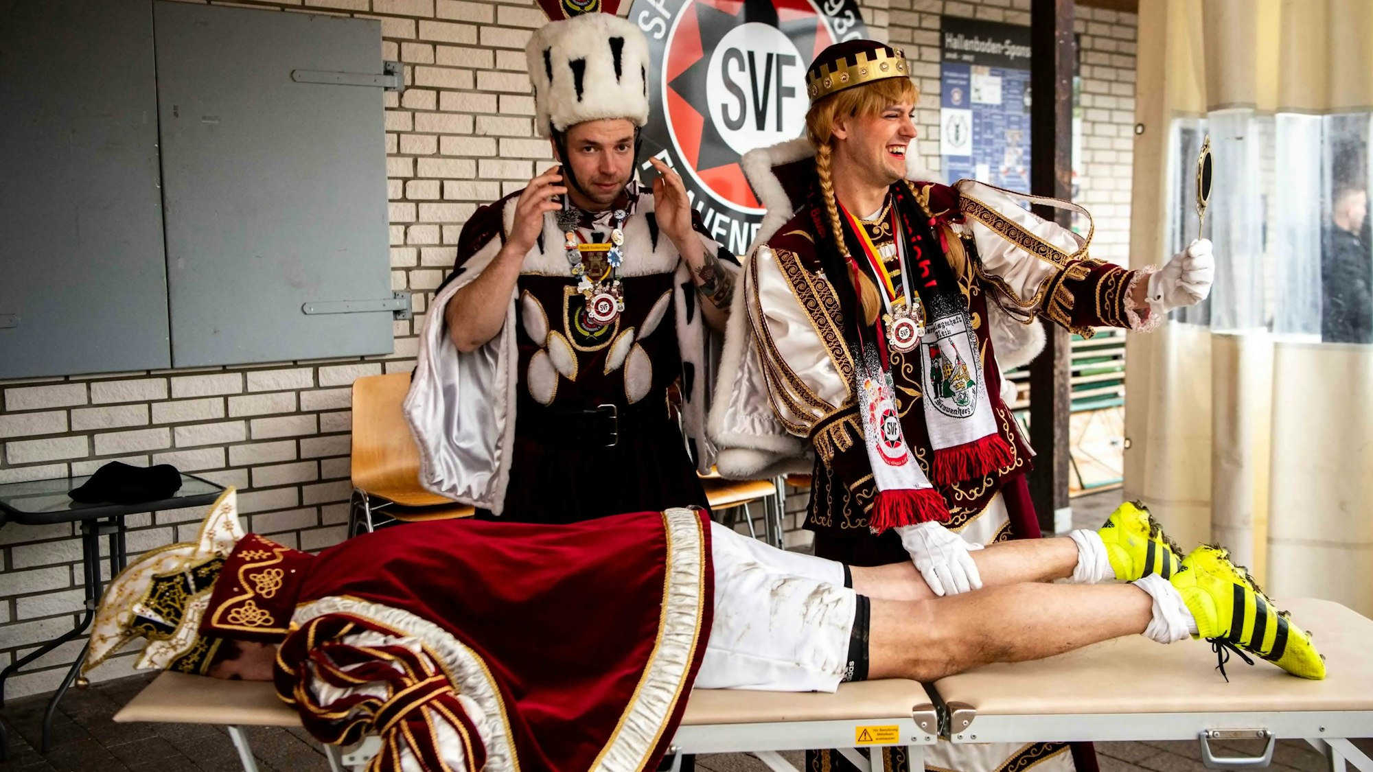 Prinz Sebastian liegt in Fußballschuhen auf der Liege, Jungfrau Olivia hat eine seiner Waden in der Hand. Bauer Charly schaut zu.