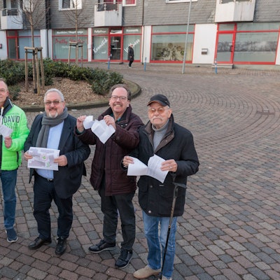 Auf der Straße „Am Marktplatz“ stehen fünf Männer und zerreißen symbolisch Papierdokumente.