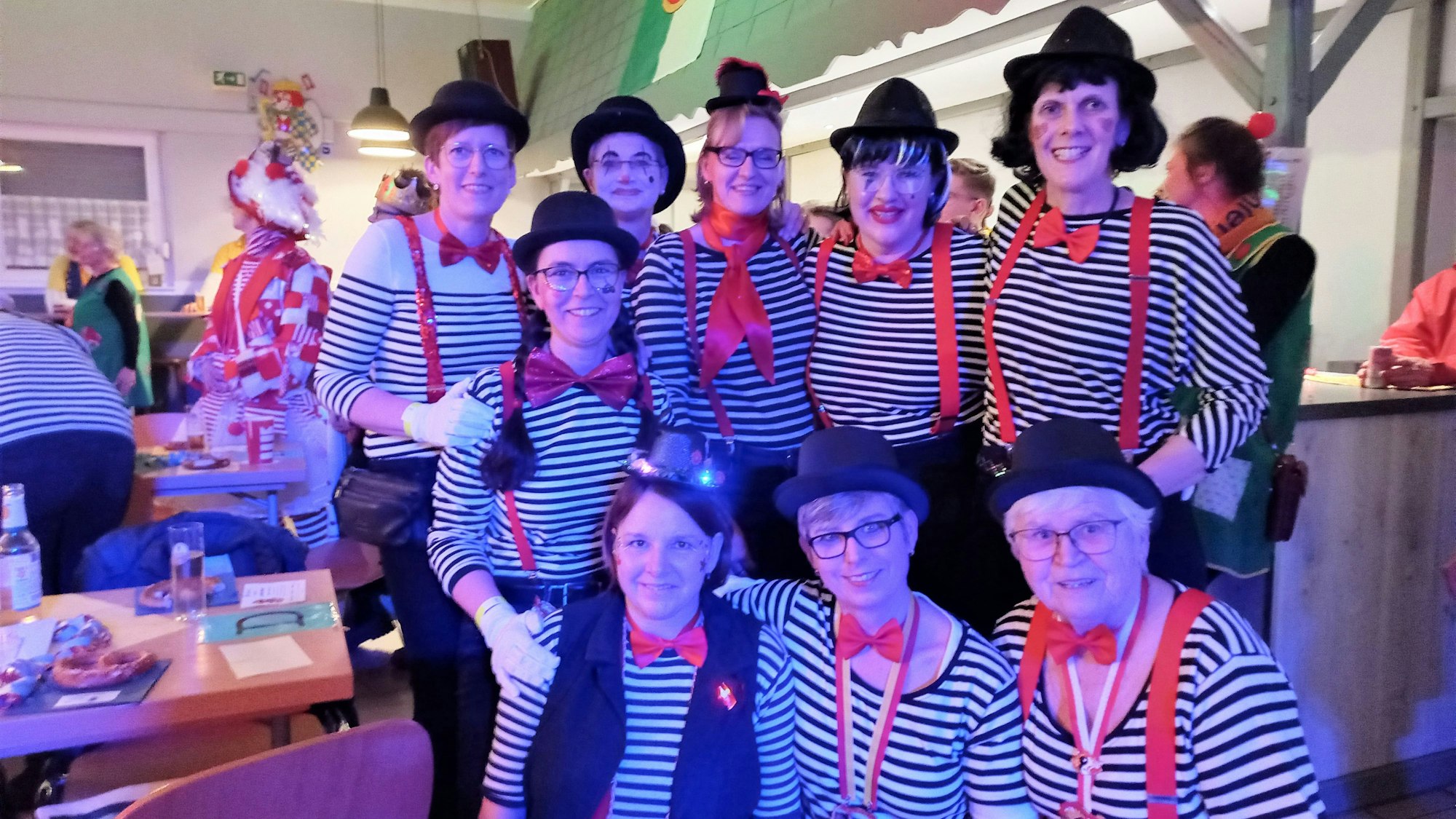 Eine Gruppe kostümierter Frauen steht zusammen. Sie alle tragen schwarz-weiß gestreifte Pullover, Hüte sowie rote Fliegen und rote Hosenträger.