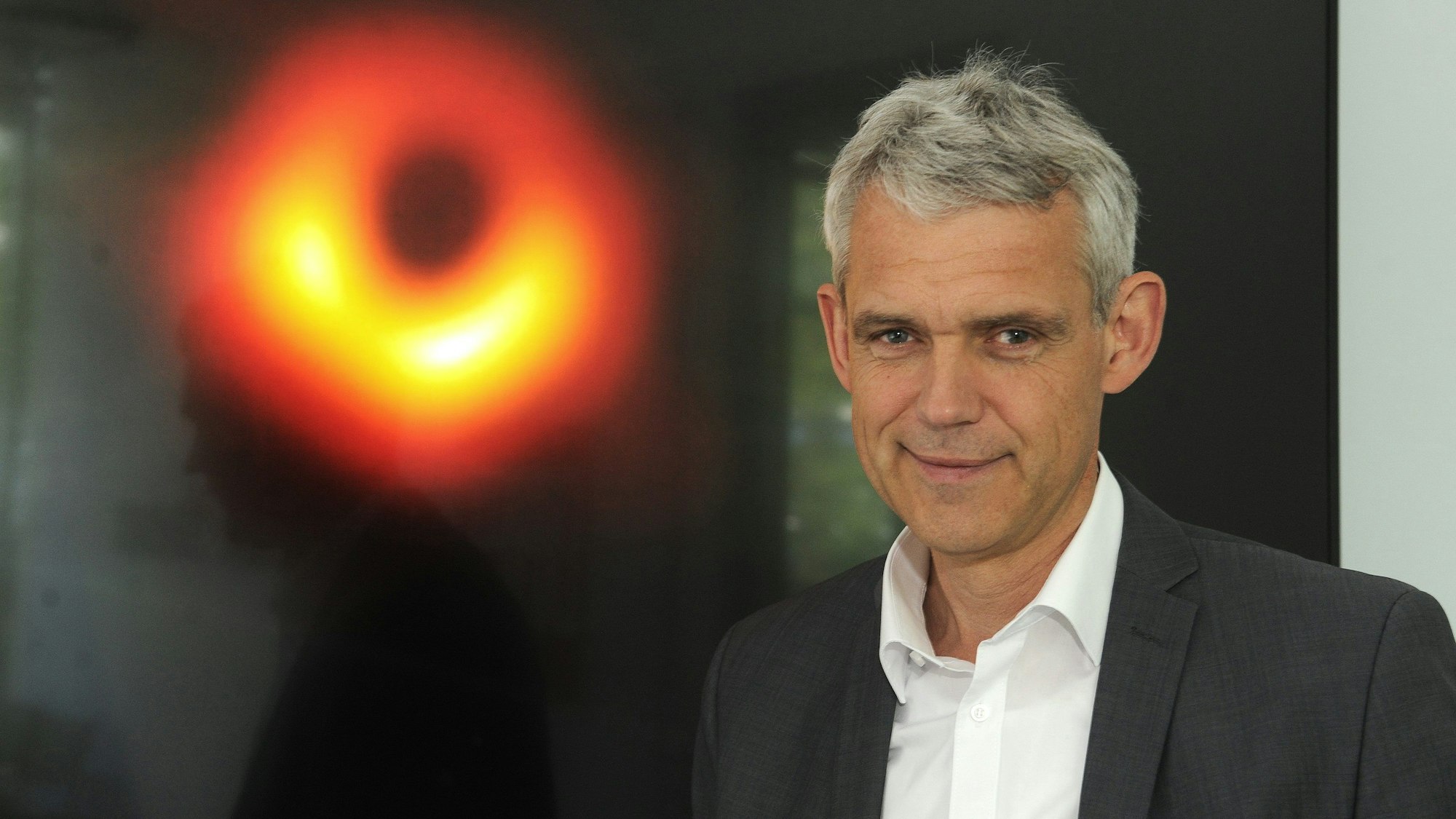Prof. Dr. Heino Falcke schaut in die Kamera. Im Hintergrund ist eine Abbildung eines schwarzen Lochs zu sehen.
