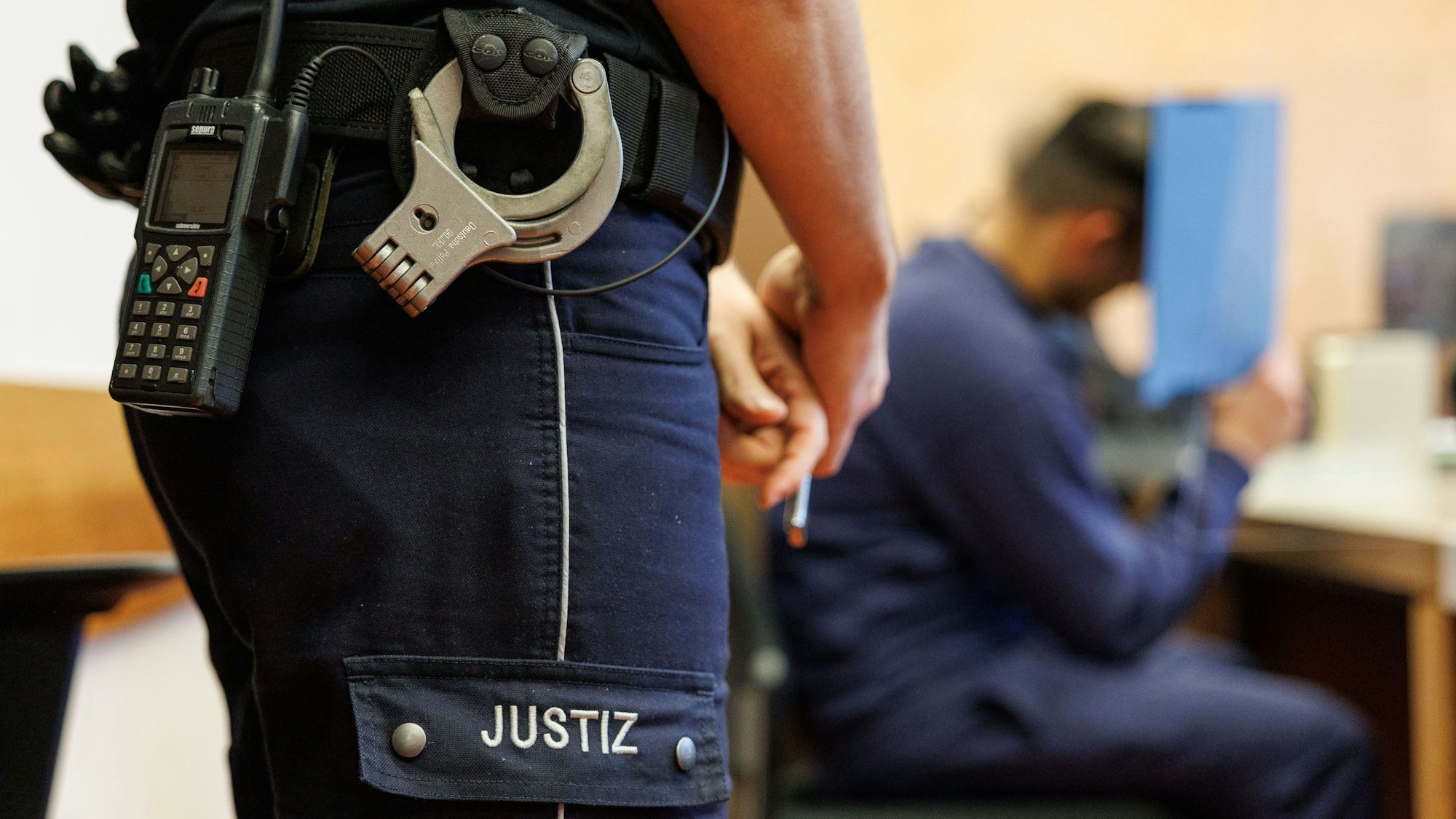 Ein Justizbeamter, mit Funkgerät und Handschellen am Hosenbund, steht neben einem Angeklagten in einem Saal im Landgericht. Der Verurteilte hat einen 39-Jährigen im Schlaf mit einer Axt getötet und anschließend seine Ex-Freundin vergewaltigt.